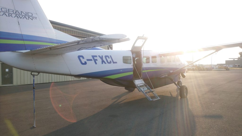 A white single engine aircraft with green and blue livery on an airport tarmac bathed in late afternoon sunshine