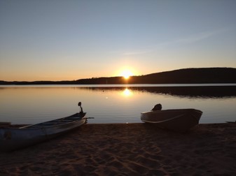 Sun setting behind a hill and reflecting into a still lake. There is a sandy beach and two boats in the foreground