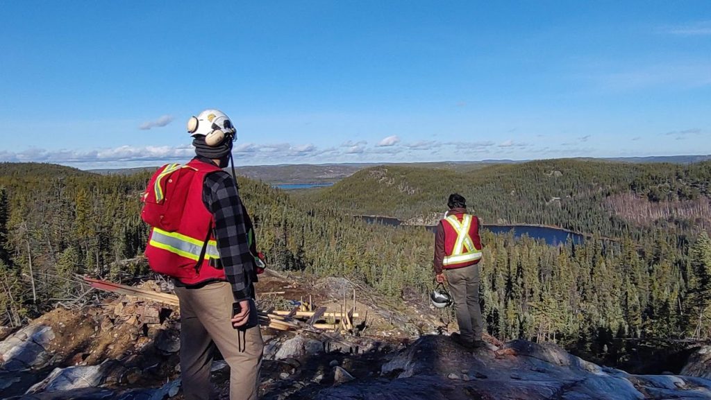 Two people standing on top of a hill overlooking a lake. Both people are wearing red cruiser vests and have their backs to the camera