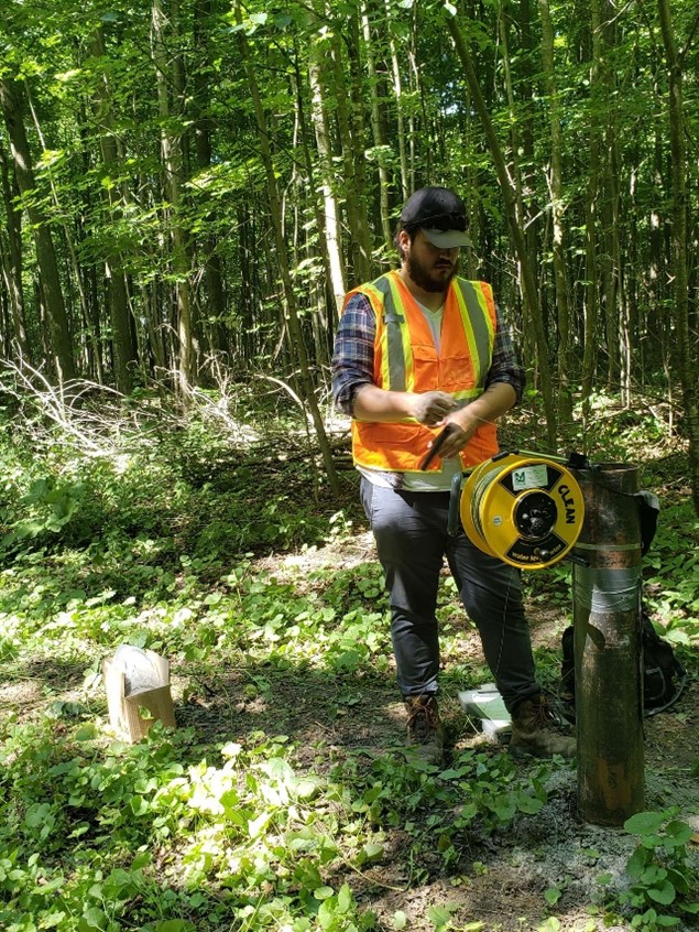 A man in a forest standing next to a well casing measuring the depth to water table using a depth tape on a yellow reel. The man is wearing a baseball cap, blue plaid shirt and an orange safety vest. He is surrounded by small trees and shrubs. 