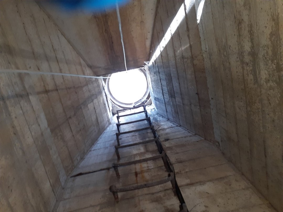 The view looking up a concrete shaft. There is a metal ladder on one wall of the shaft. Light is shining down from a circular opening.