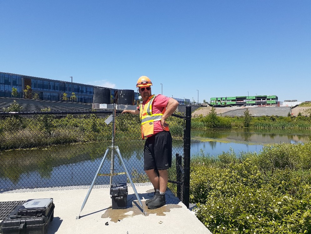 A man posing on a concrete pad on the edge of a storm water pond. He is wearing a red shirt, orange reflective vest, black shorts and work boots. He is standing next to a tripod.