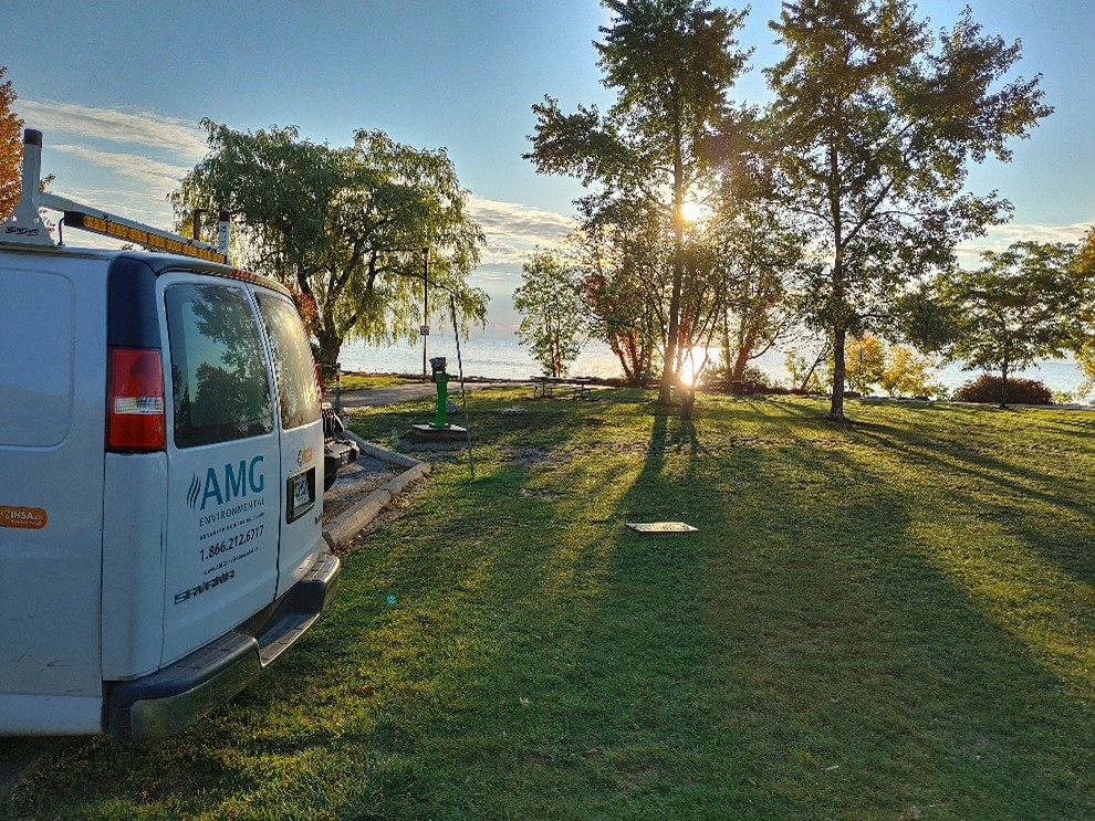 Sun rising over a lake and shining through the trees. The sky is mostly clear with a few streaky cloudes. A white work truck is partly visible on the left.