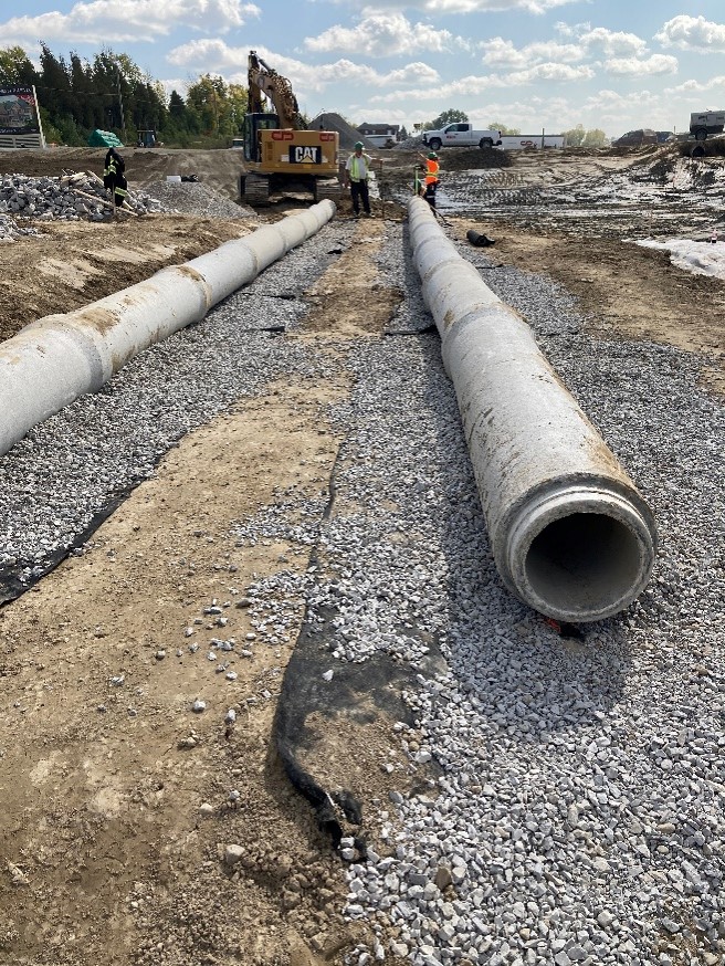 Large diameter concrete pipes lying on gravel-covered geotextile. There is a pickup truck, an excavator and three people in the background. 