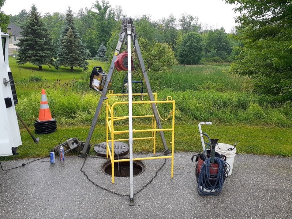 A yellow safety cage and large metal tripod set up over an open maintenance hole in a rural area. There are buckets of tools and a stack of safety cones.